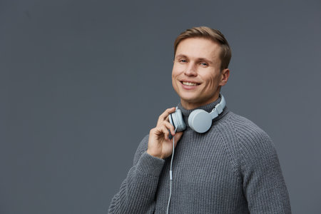 smiling young caucasian man wear gray sweater hold headphones on neck wireless communication mental...