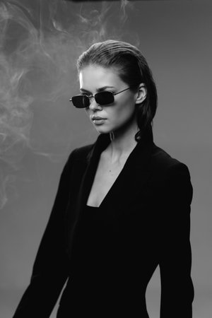 Black and white portrait of confident woman wearing sunglasses and stylish black outfit with smoke...