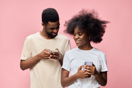 Happy young couple texting on smartphones against a soft pink background, showcasing modern...