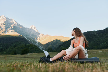 A relaxed outdoor scene shows a person sitting on the grass with a laptop, gazing toward distant...