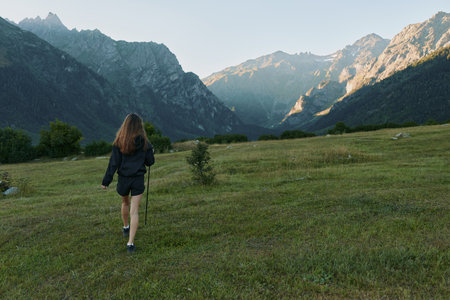 A lone hiker walks across an open meadow toward rugged mountains, creating a mood of adventure,...