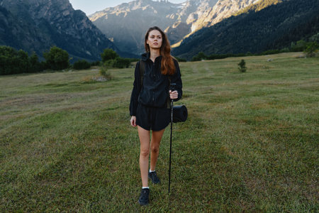 A solo hiker stands in a grassy meadow with distant mountains behind her. She wears a dark jacket...