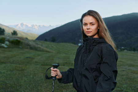 A focused adventurer stands in a green valley with a trekking pole, wearing a dark jacket. She gazes...