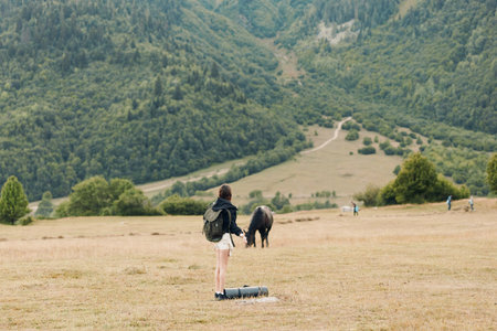 A solitary traveler stands in a wide meadow, backpack resting by their side while a horse grazes in...