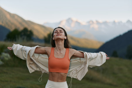 A carefree woman stands in a scenic mountain meadow with arms outstretched, wearing an orange sports...