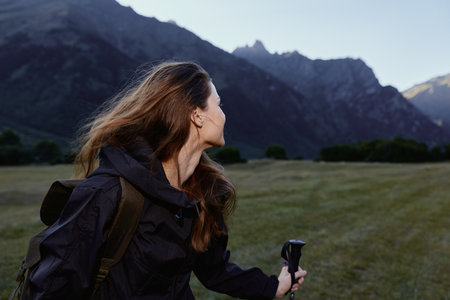 A focused hiker with a backpack stands in an open meadow, looking toward distant rugged peaks. She...