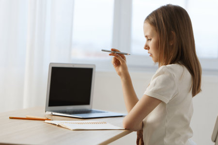 Thoughtful young girl studying at home with a laptop, notebook, and pencil, reflecting concentration...
