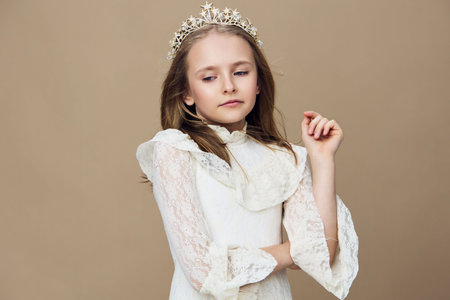 Young girl with a crown, wearing a white lace dress, exuding elegance and confidence, against a neutral beige backgroundの写真素材