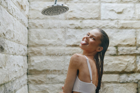 smiling woman enjoying outdoor shower under rain with wet hair by stone wall, carefree and playful...