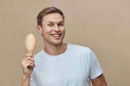 Smiling happy young caucasian man holding hairbrush casual white t-shirt natural beige background personal care grooming conceptの写真素材