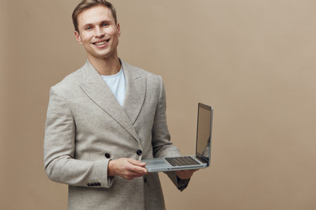 smiling young man holding laptop wearing jacket standing isolated on beige background remote work...