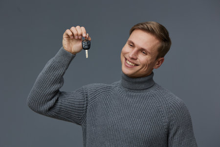 Smiling young man with short brown hair wearing a gray turtleneck sweater holding car key on gray...