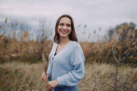 Smiling young woman in a cozy sweater holds wild grass in a windswept field, captured with natural light, relaxed posture and a genuine smile to convey authenticity and honest everyday life.の写真素材