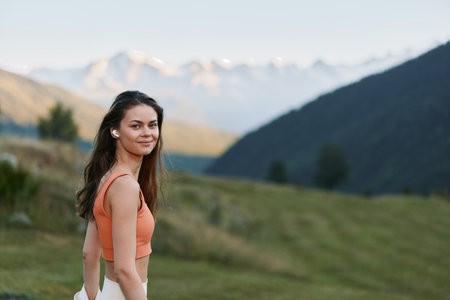 A cheerful woman stands outdoors in a scenic mountain landscape. She smiles warmly, wearing a coral top, exuding confidence and a relaxed, adventurous vibe.の写真素材