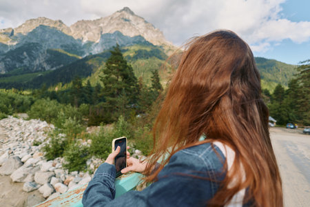 A woman with long hair stands outdoors, using a smartphone against a mountainous landscape. The...