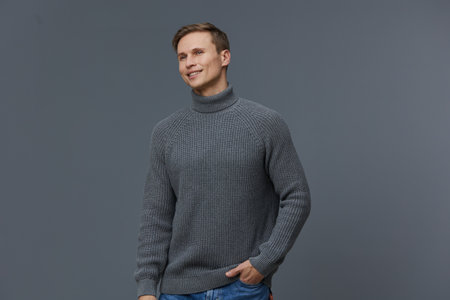 Smiling man wearing gray knitted turtleneck sweater on plain gray background, casual fashion,...