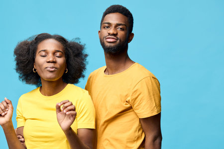 Joyful young African couple dancing together in matching yellow t shirts against a vibrant blue...