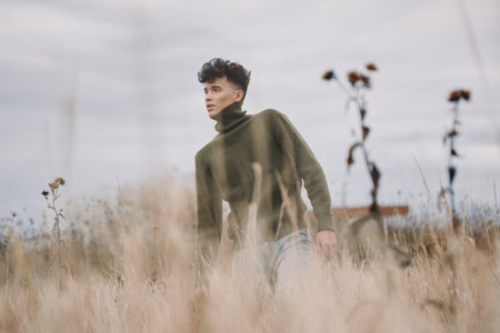 A young man in a green knit turtleneck walks through tall dry grass, captured in a candid, authentic portrait style conveying genuine presence, natural texture, subtle movement and introspective mood.の写真素材
