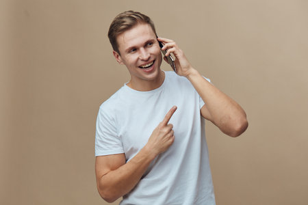 smiling young man talking phone pointing finger wearing white t-shirt isolated light brown...