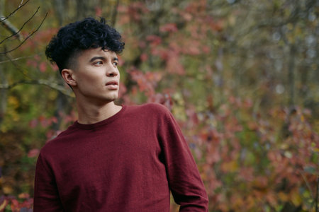 An authentic portrait of a young man in a burgundy sweater stands amid autumn foliage, conveying veracity through natural posture, thoughtful gaze, and genuine, unposed mood.の写真素材