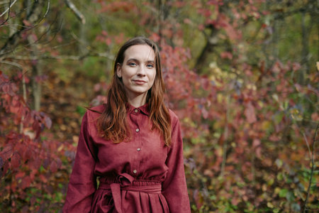 Authentic portrait of a young woman standing in an autumn wood, wearing a burgundy coat, calm...
