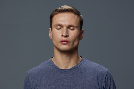 Calm young man with closed eyes and relaxed expression wears casual blue sweater against neutral...