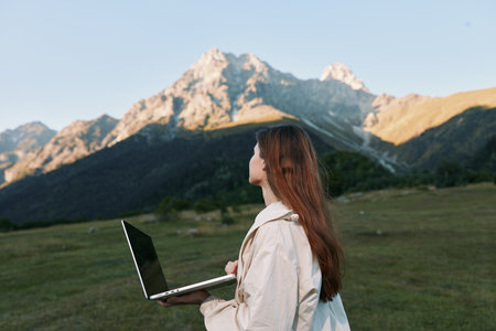 A cheerful woman stands outdoors with a laptop in a scenic mountainous landscape. She wears a light...