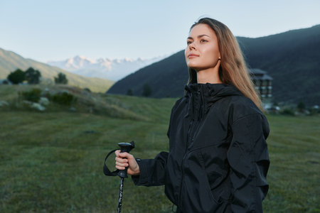 A female hiker stands in a meadow, gripping a trekking pole and gazing into the distance. The...