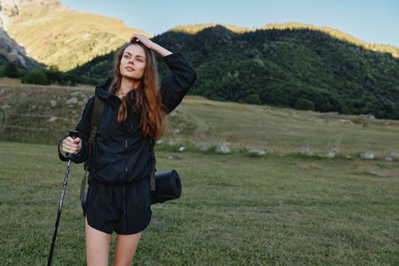 A cheerful outdoor enthusiast stands in a grassy field with a trekking pole, wearing a dark jacket...