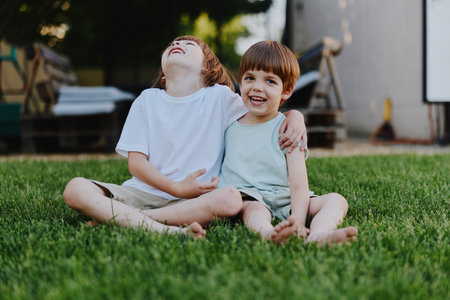 Joyful boys sitting on grass, laughing together, showcasing friendship and childhood innocence in a...