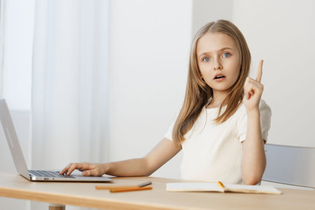 Thoughtful girl with long hair sitting at a desk, raising her finger in a moment of inspiration, using a laptop and notebook, focused on her work and ideasの写真素材