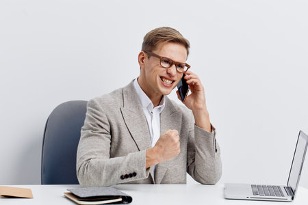 Professional man in glasses and suit smiling while talking on smartphone in modern office environment, sitting at desk with laptop and notepad, expressing success and confidence, business communication concept.の写真素材
