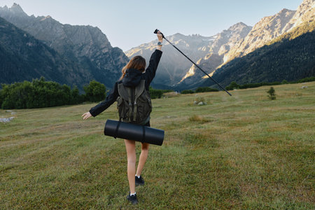 A focused hiker walks across a grassy meadow toward dramatic mountains, carrying a backpack and pole, enjoying outdoor adventure in wide open space under clear blue skies.の写真素材