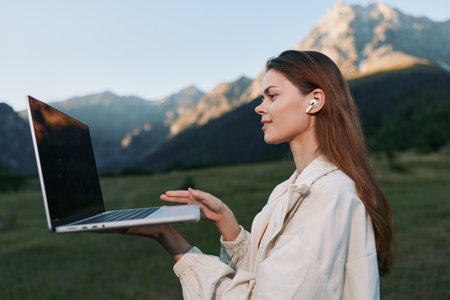 A focused business professional stands outdoors in a scenic mountain setting, balancing a laptop in...