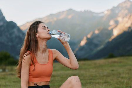 A young woman sits on the grass in a mountain landscape, drinking water from a bottle after a...