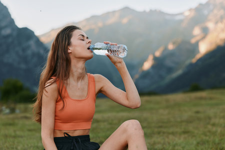 A woman in an orange sports top drinks water from a bottle in a scenic mountain meadow. She sits on...