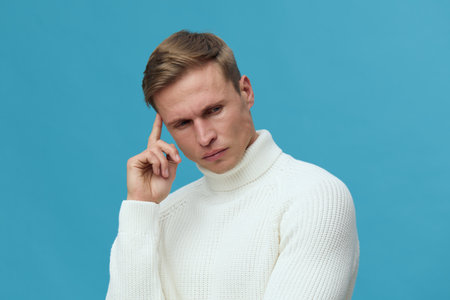Close-up of concentrated young Caucasian man with blond hair wearing white turtleneck sweater,...