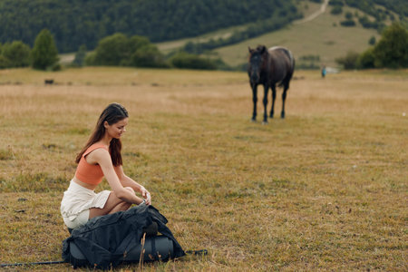 A young woman sits on a grassy field with a backpack, preparing gear outdoors. She wears casual clothes and appears calm, as a horse grazes in the distance against a tranquil landscape.の写真素材