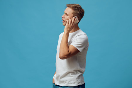 Man talking on mobile phone isolated on plain solid blue background. Studio portrait of a young casual man with short hair wearing white t-shirt using cellphone. People lifestyle concept, communication, technology.の写真素材
