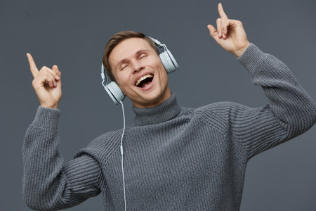 Joyful man with headphones expressing happiness, dancing and enjoying music in a casual gray sweater, against a plain gray background. People lifestyle concept, positive emotion, leisure activity, relaxation, music enthusiast.の写真素材