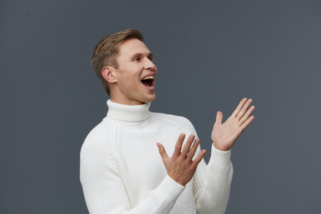 A joyful young man with light brown hair, wearing a cozy white turtleneck sweater, stands against a plain gray background. He is smiling broadly and gesturing with his hands, expressing excitement and happiness in this studio portrait. People lifestyle conceptの写真素材