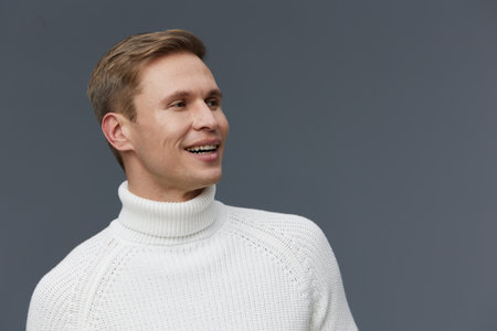 Smiling man wearing white turtleneck sweater looking to the side on plain gray background studio portrait. People lifestyle concept, positive expression, casual style, warm clothing, relaxed mood, horizontal composition.の写真素材