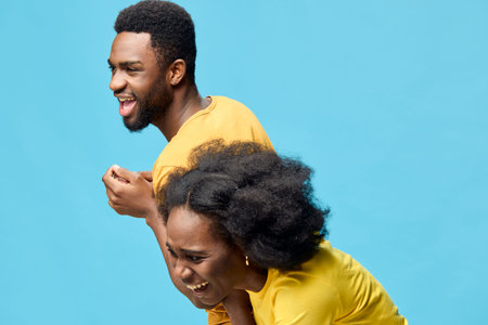 Joyful young African couple laughing together against a bright blue background, wearing matching...