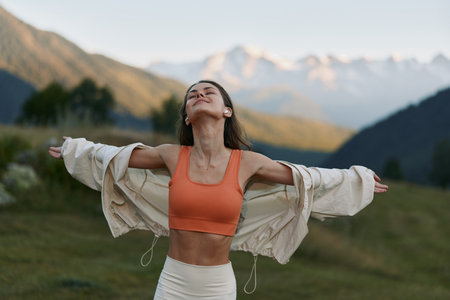 A carefree woman stands outdoors with arms spread wide, wearing an orange sports bra and a light robe. She breathes fresh air and savors freedom amid majestic mountain scenery, inspiring vitality.の写真素材