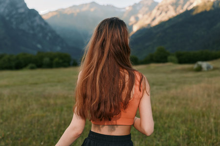 A woman stands in a sunlit meadow, facing distant mountains. Her long hair falls over her back as she gazes across the open landscape, evoking calm, serenity, and a sense of freedom in nature.の写真素材