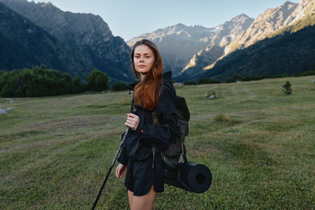 A young woman hiker stands in a wide alpine meadow with towering mountains in the background. She carries a backpack and trekking pole, dressed in outdoor gear, looking ahead with confidence andの写真素材