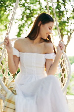 Young woman in a white dress smiling gently while sitting on a swing, surrounded by green trees and soft sunlight, conveying tranquility and femininityの写真素材
