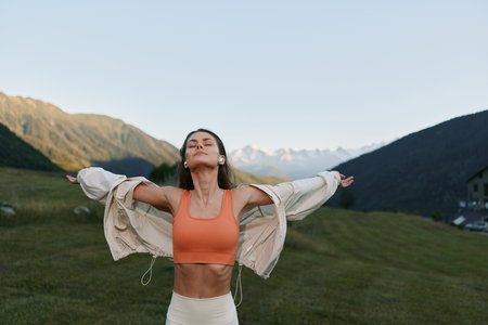 A carefree woman stands outdoors with arms wide, wearing athletic gear in a scenic mountain meadow. She looks joyful and liberated, savoring fresh air and wide horizons.の写真素材