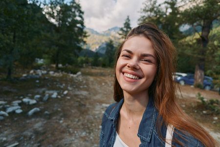 A cheerful woman stands outdoors in a natural setting, smiling warmly as sunlight glints on her denim jacket. She appears relaxed, confident, and ready for exploration.の写真素材