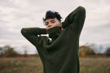 A candid portrait of a young man in a thick green sweater, exuding authenticity and quiet confidence under soft outdoor light, with a calm, natural expression and subtle mood.の写真素材